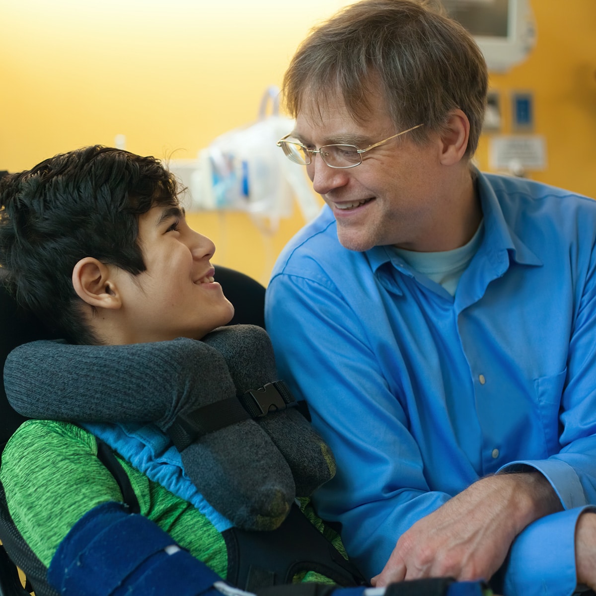 Smiling father sitting next to disabled son in wheelchair by hospital bed talking together